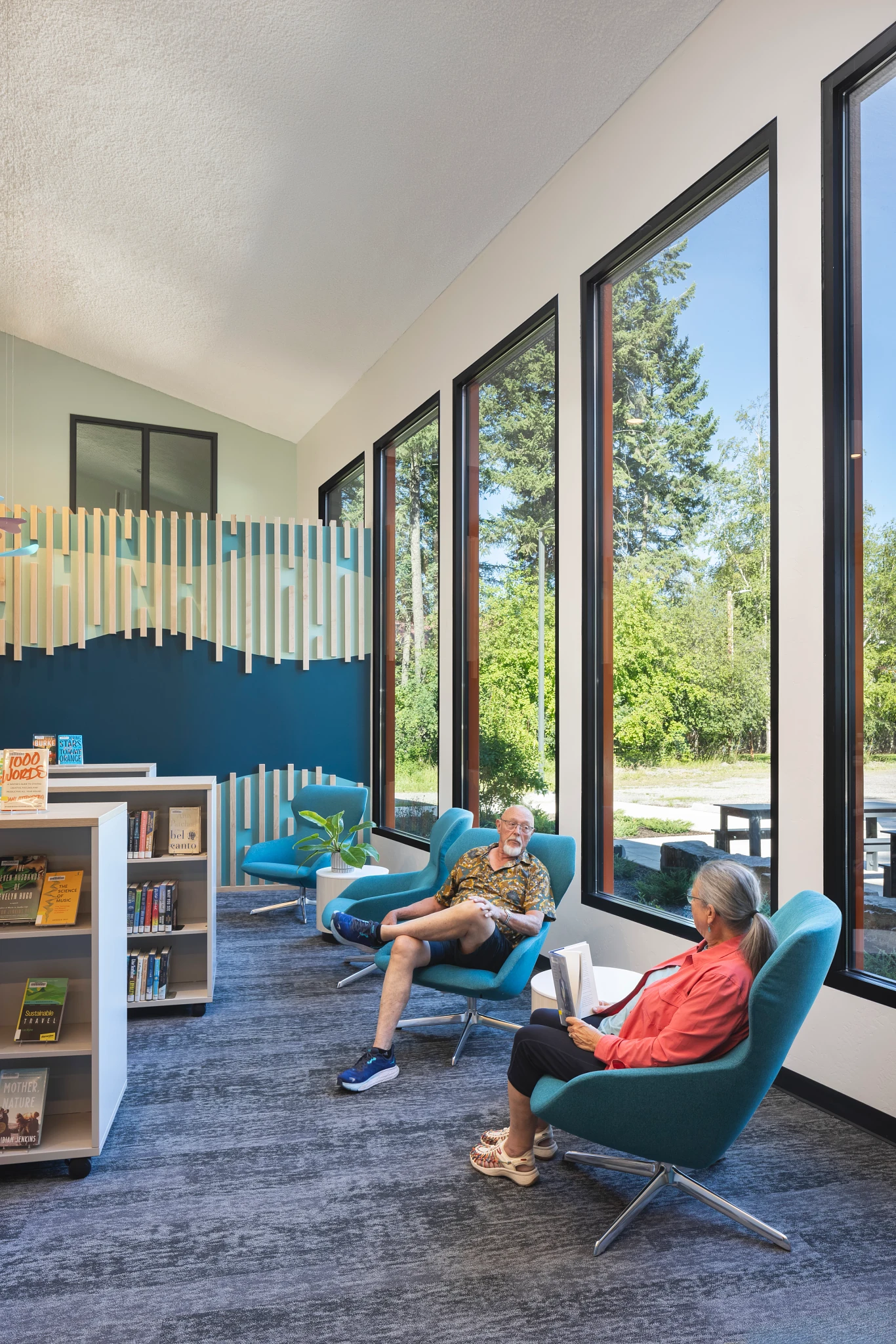 Education facility with commercial carpet tile flooring in a library setting, featuring blue and gray tones, 9x36 inch dimensions, with people reading and relaxing in teal chairs near large windows.