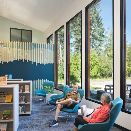 Education Facility Library with Commercial Carpet Tile Flooring in Blue and Gray Tones Education facility with commercial carpet tile flooring in a library setting, featuring blue and gray tones, 9x36 inch dimensions, with people reading and relaxing in teal chairs near large windows.