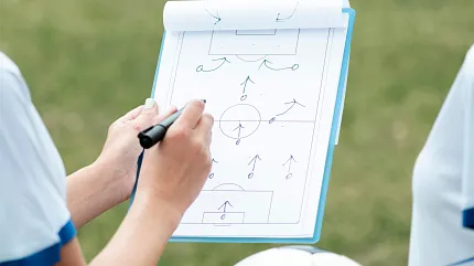 Coach drawing a soccer game plan with arrows on a clipboard during a strategy session on the field. Coach drawing a soccer game plan with arrows on a clipboard during a strategy session on the field.