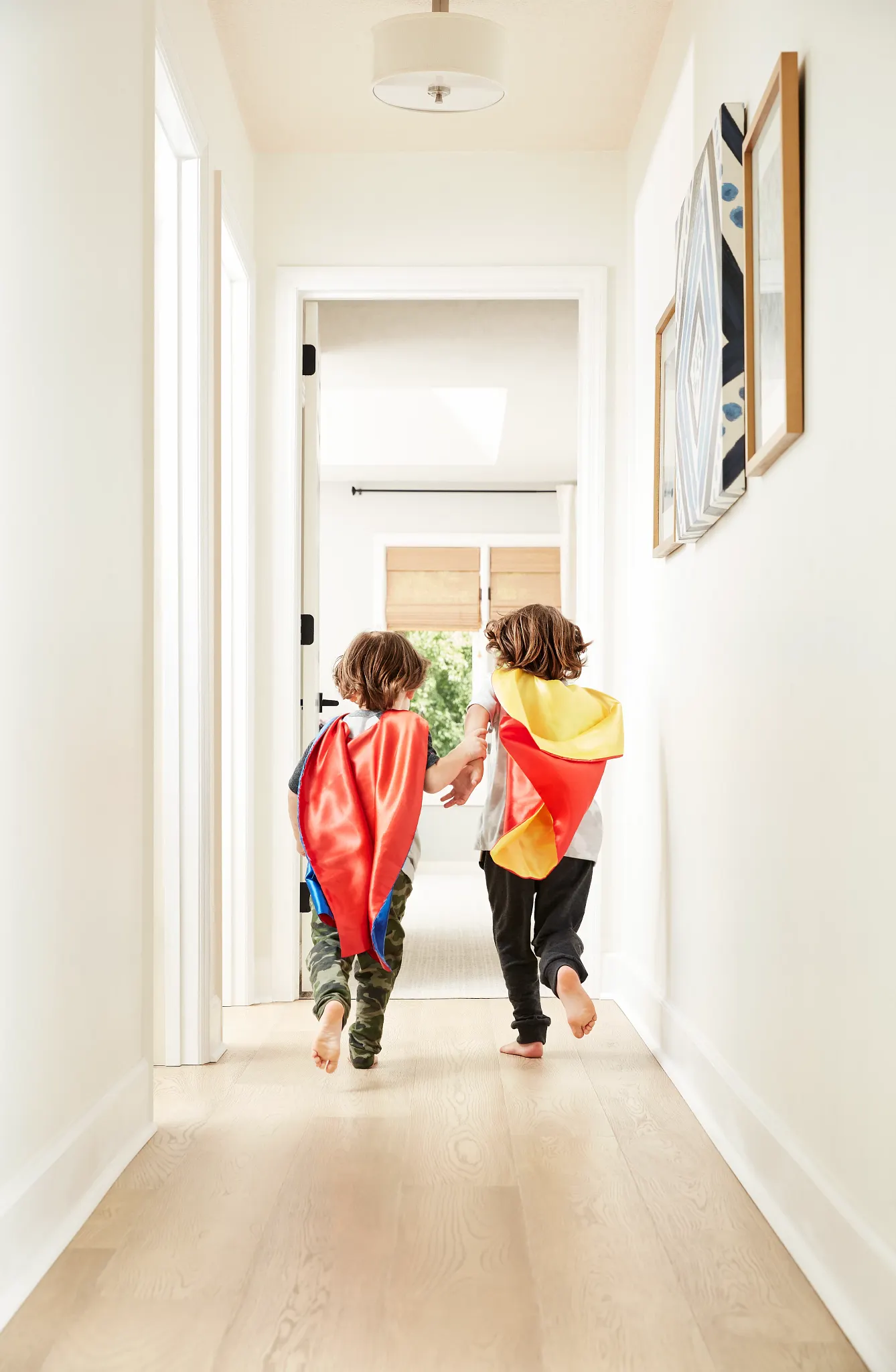 Two children wearing capes running down a hallway with light wood flooring