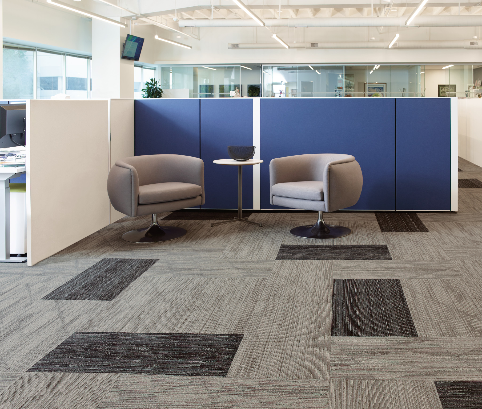 
  "Modern office space with commercial carpet tiles in shades of gray and dark gray, featuring two gray chairs and a small round table against a blue partition"
)