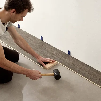 Easy Installation of Wood Look Luxury Vinyl Plank Flooring. Man installing luxury vinyl plank flooring with a rubber mallet and spacer blocks, showcasing easy installation and wood look design.