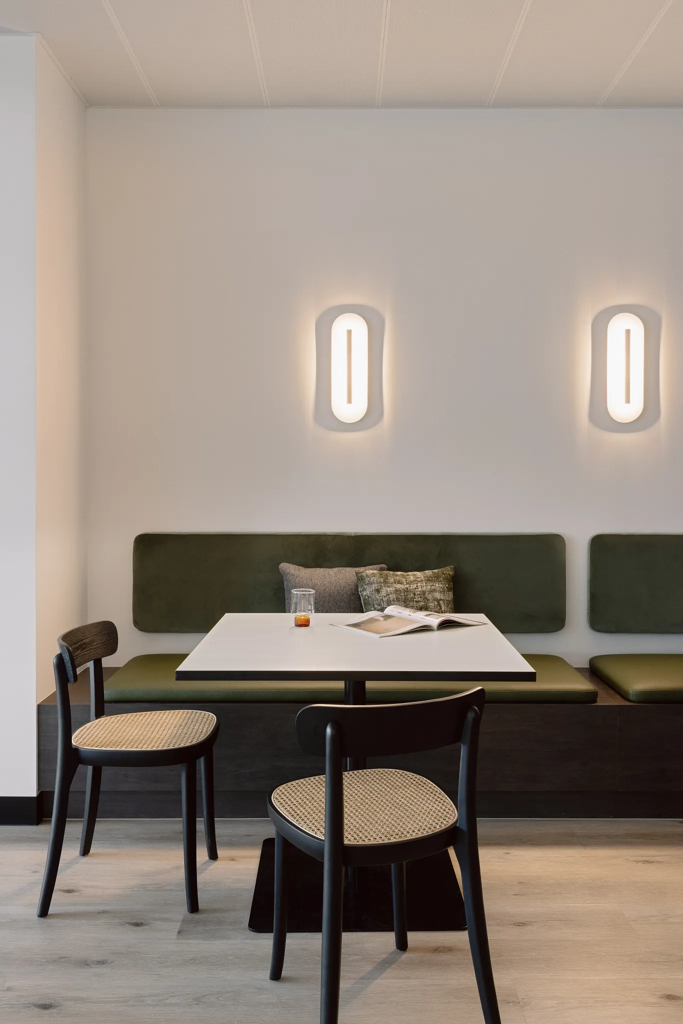 Modern seating area with green cushioned bench, black chairs, and light wood flooring in a hospitality setting
