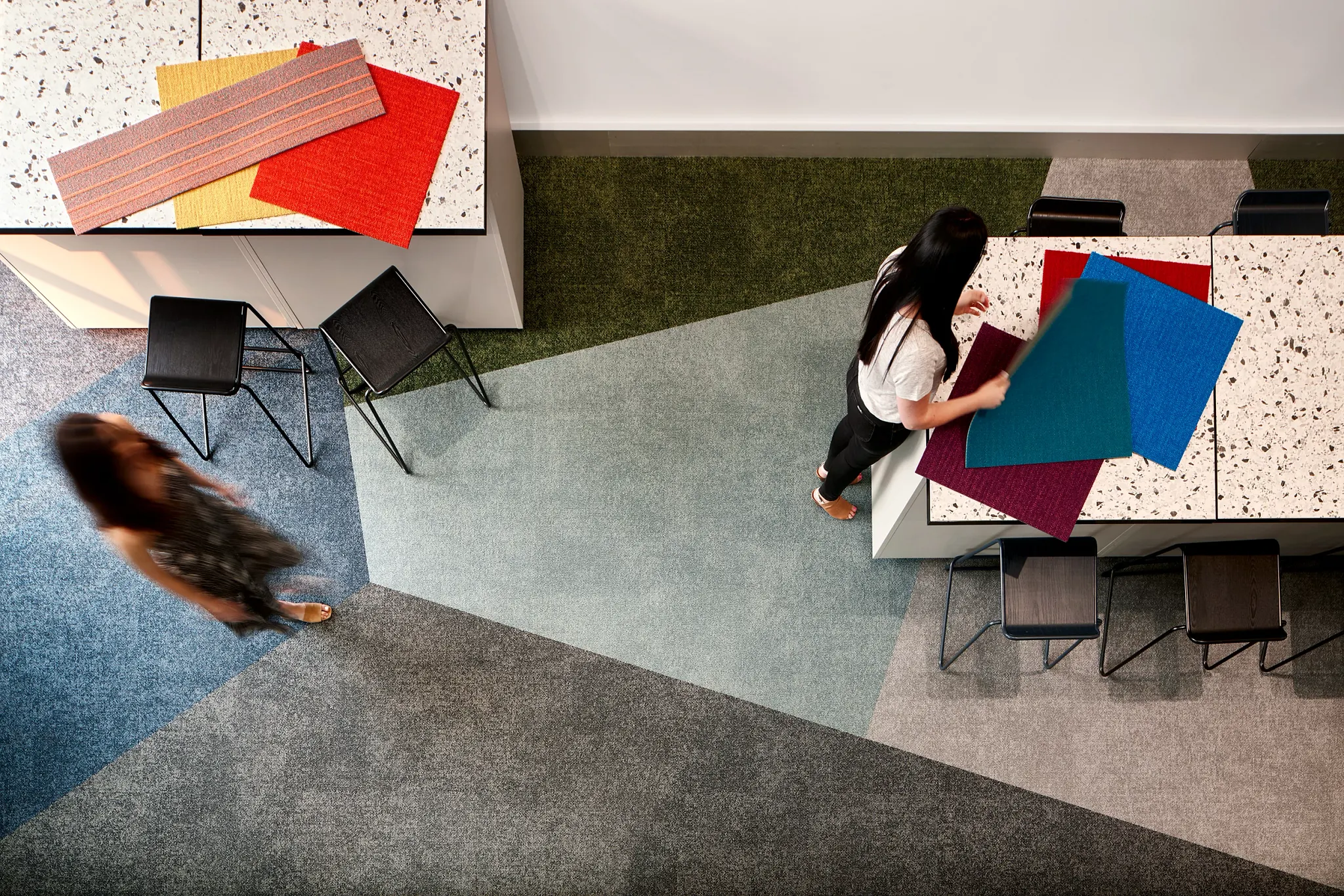 Overhead view of a modern education facility with geometric patterned commercial carpet tile in shades of blue, green, and gray, featuring two women interacting with colorful fabric swatches on a terrazzo table