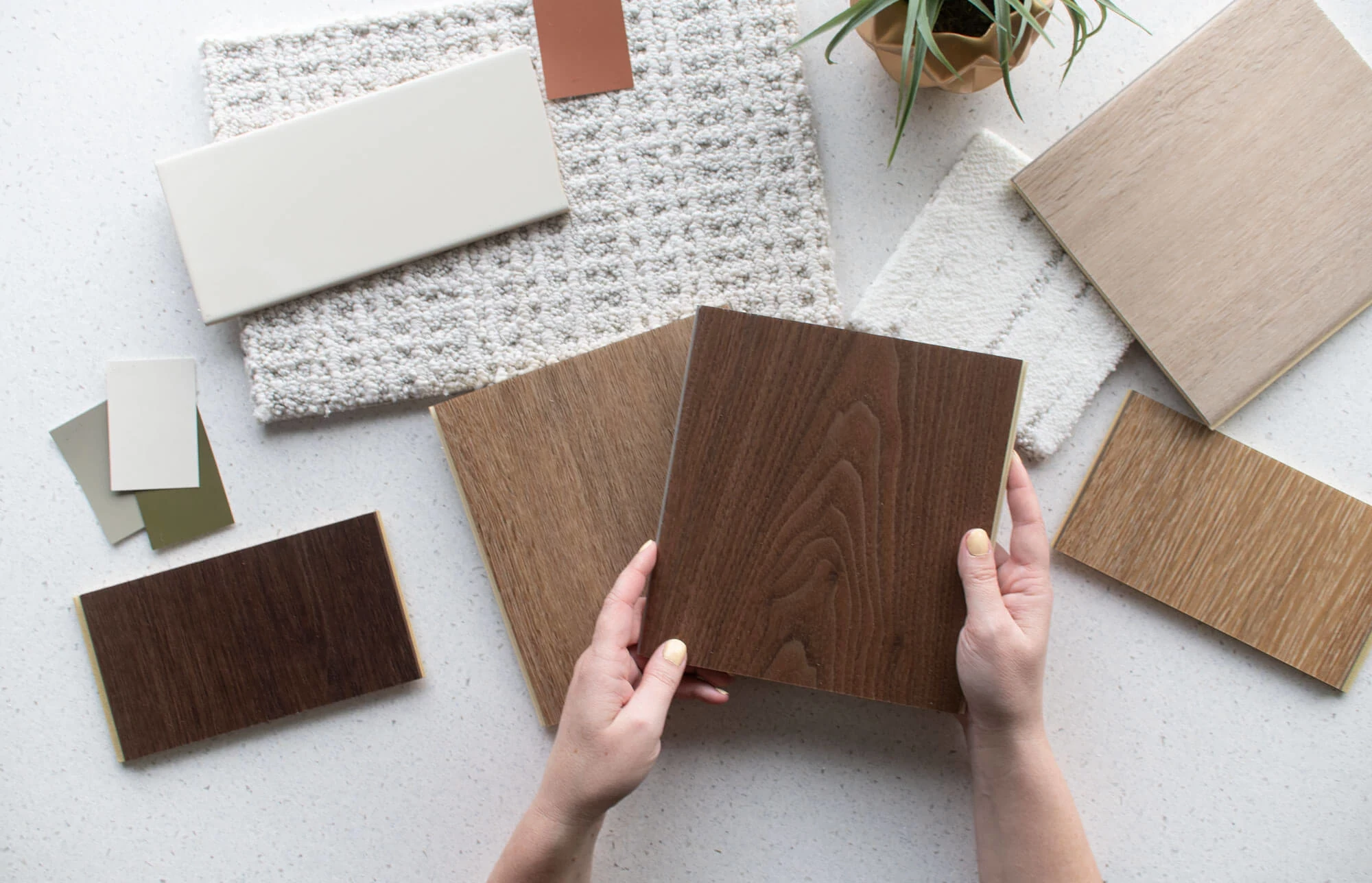 Hands holding a wood look vinyl flooring swatch surrounded by various flooring samples in shades of brown, beige, and cream