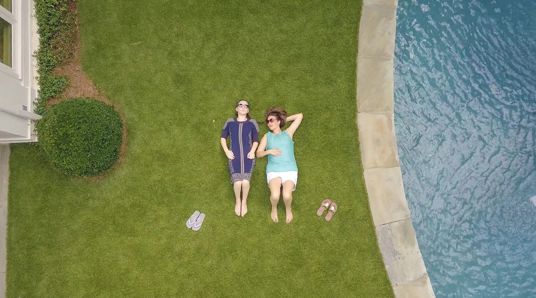 Aerial view of two people relaxing on a lush green lawn next to a swimming pool