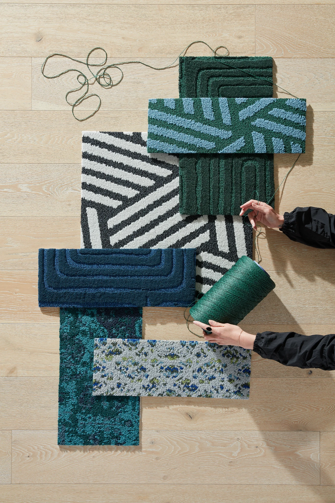 Assorted commercial carpet tile swatches in green, blue, and black patterns on light wood flooring with hands holding green yarn