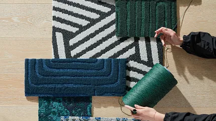 Commercial Carpet Tile Swatches on Light Wood Flooring Assorted commercial carpet tile swatches in green, blue, and black patterns on light wood flooring with hands holding green yarn