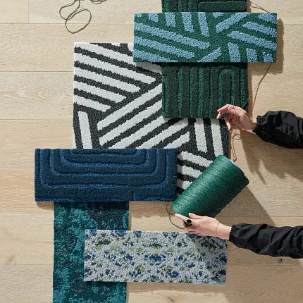Commercial Carpet Tile Swatches on Light Wood Flooring Assorted commercial carpet tile swatches in green, blue, and black patterns on light wood flooring with hands holding green yarn