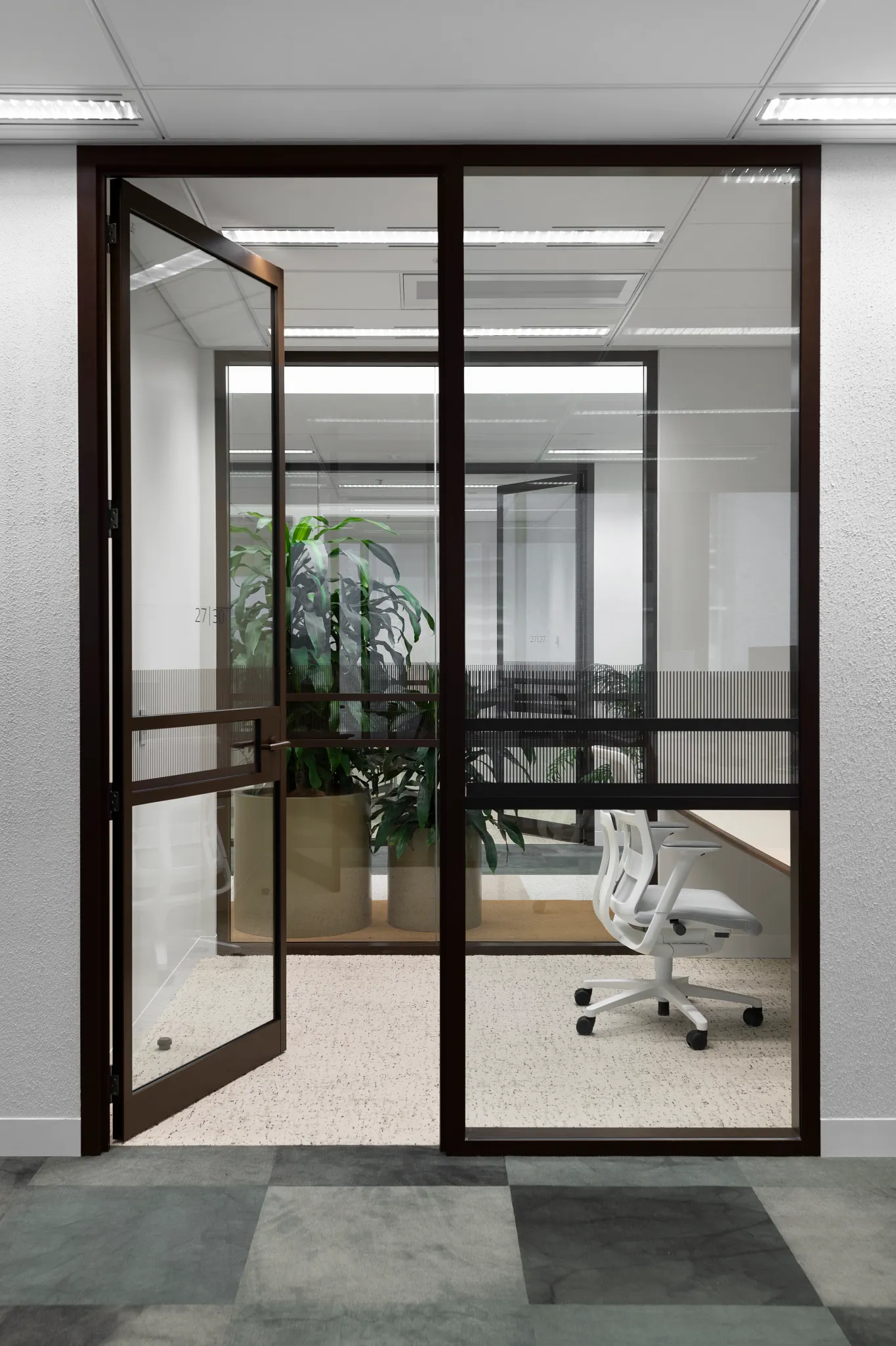 Modern office space with glass partitions, white office chair, and green potted plants, featuring a mix of light and dark gray commercial carpet tiles