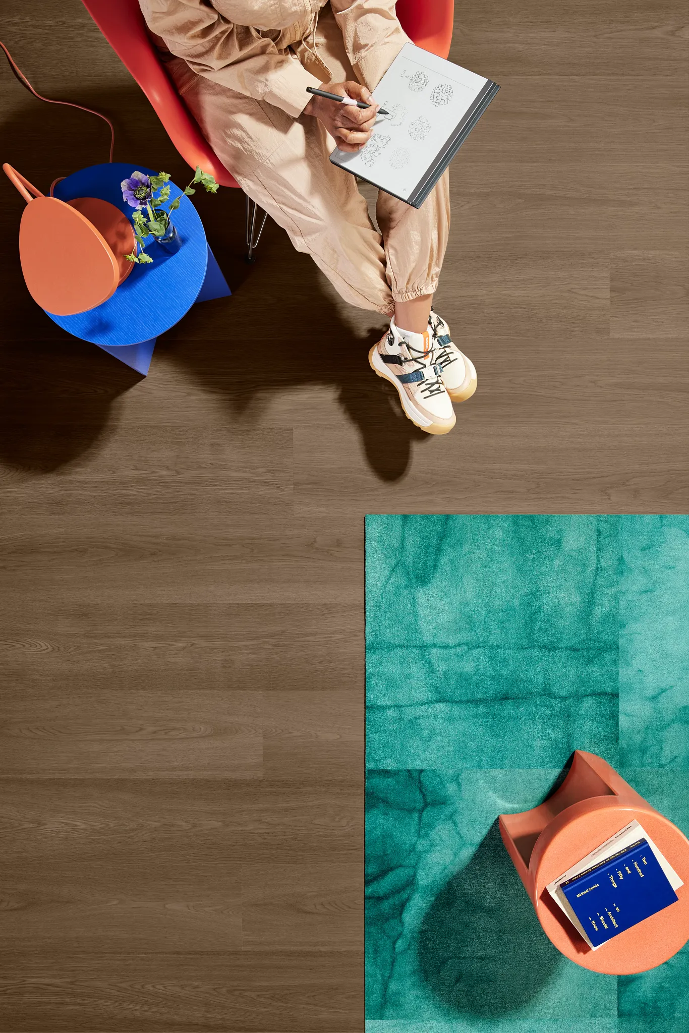 Overhead view of a modern workspace featuring Heavy Commercial PVC-Free EcoWorx Resilient flooring in a warm brown tone, with a person sketching in a notebook, a vibrant blue and orange side table, an