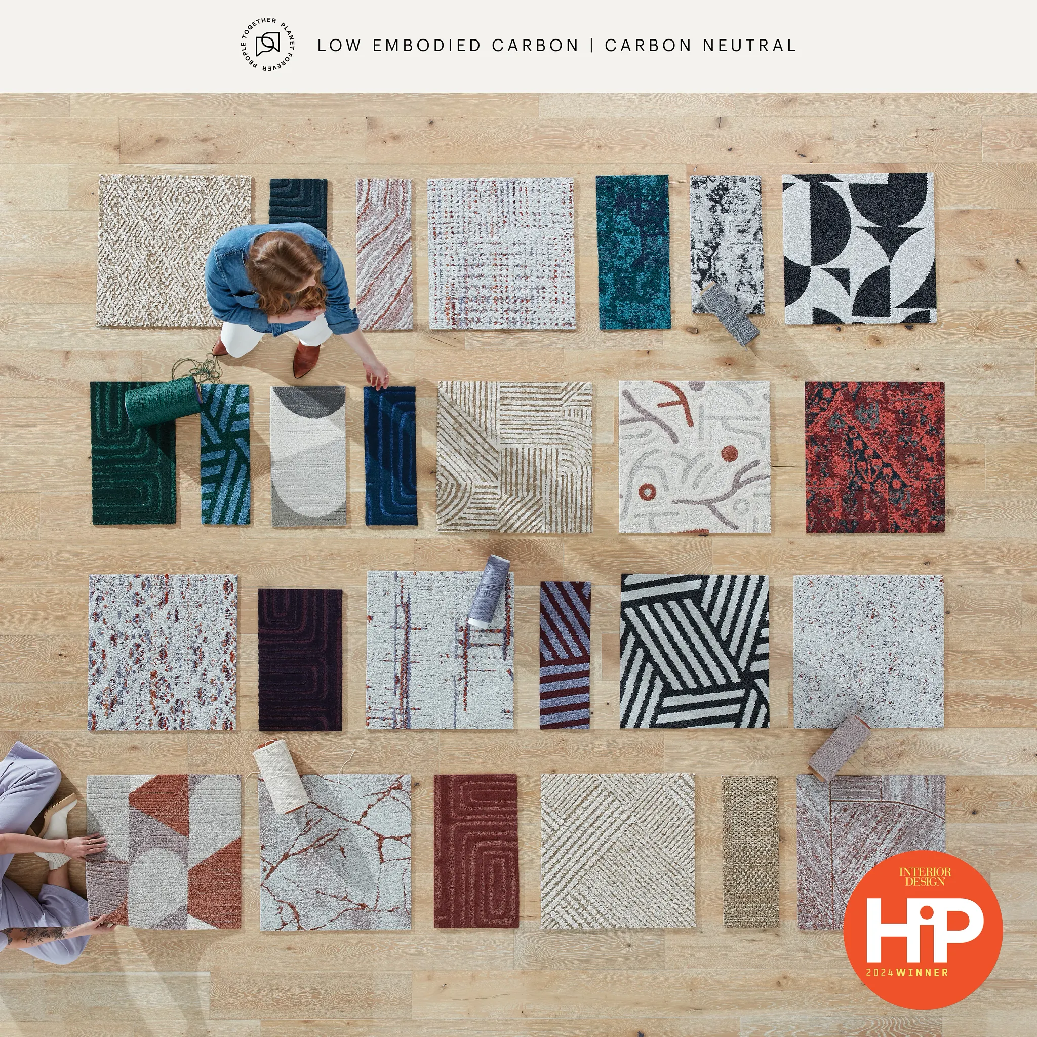Overhead view of diverse commercial carpet tile swatches in various colors and patterns, including beige, blue, red, and black, with two people arranging them on a wooden floor