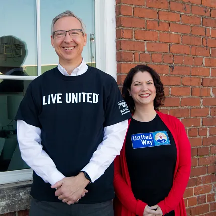 People in "Live United" and "United Way" shirts in front of a brick building. Two people standing in front of a brick wall and window, wearing "Live United" and "United Way" shirts.