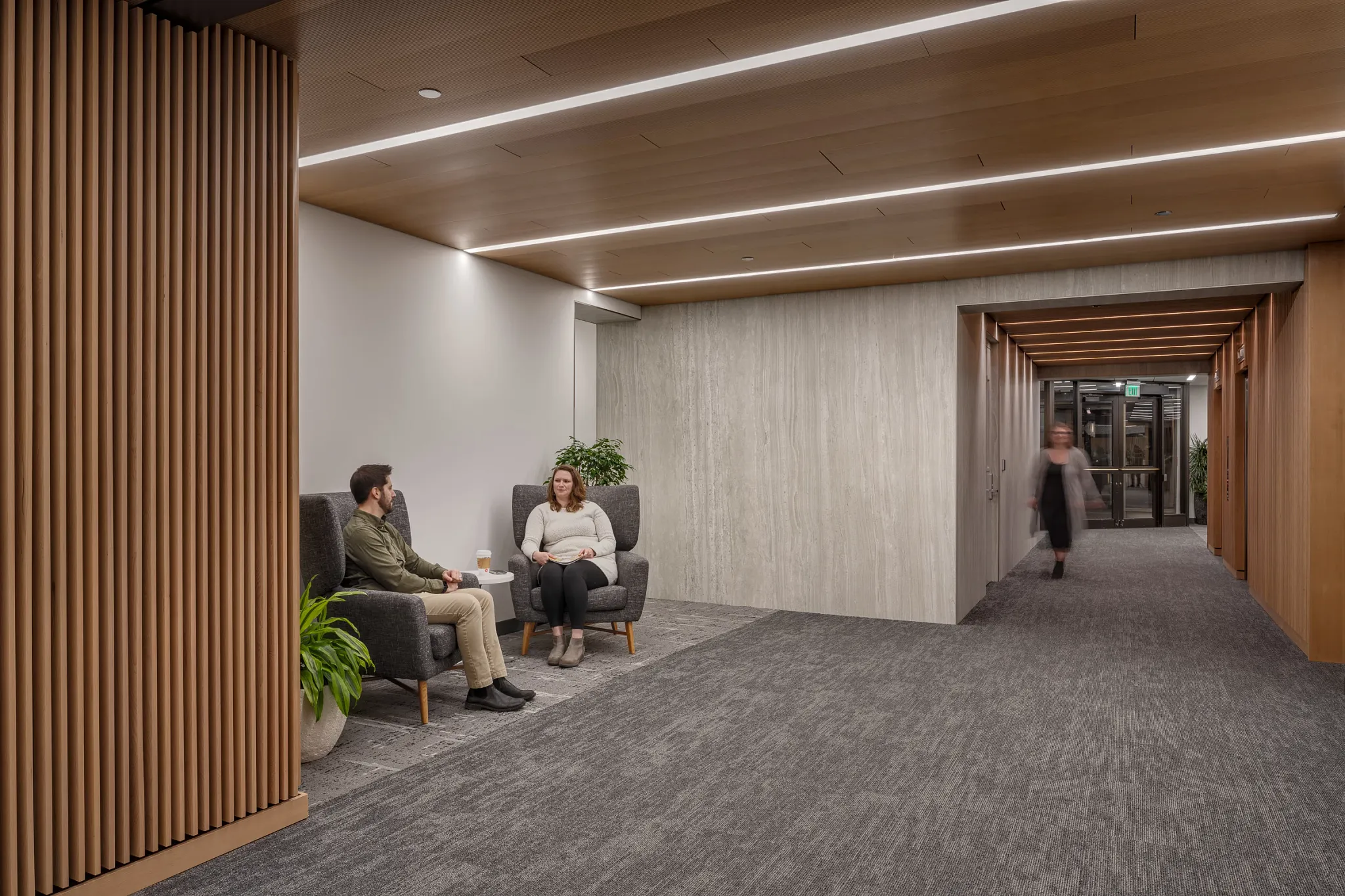 Modern workplace hallway with gray commercial carpet tile flooring, featuring two people seated in armchairs and a person walking