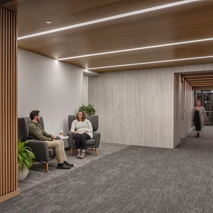 Contemporary workplace hallway with gray commercial carpet tile flooring and seating area Modern workplace hallway with gray commercial carpet tile flooring, featuring two people seated in armchairs and a person walking