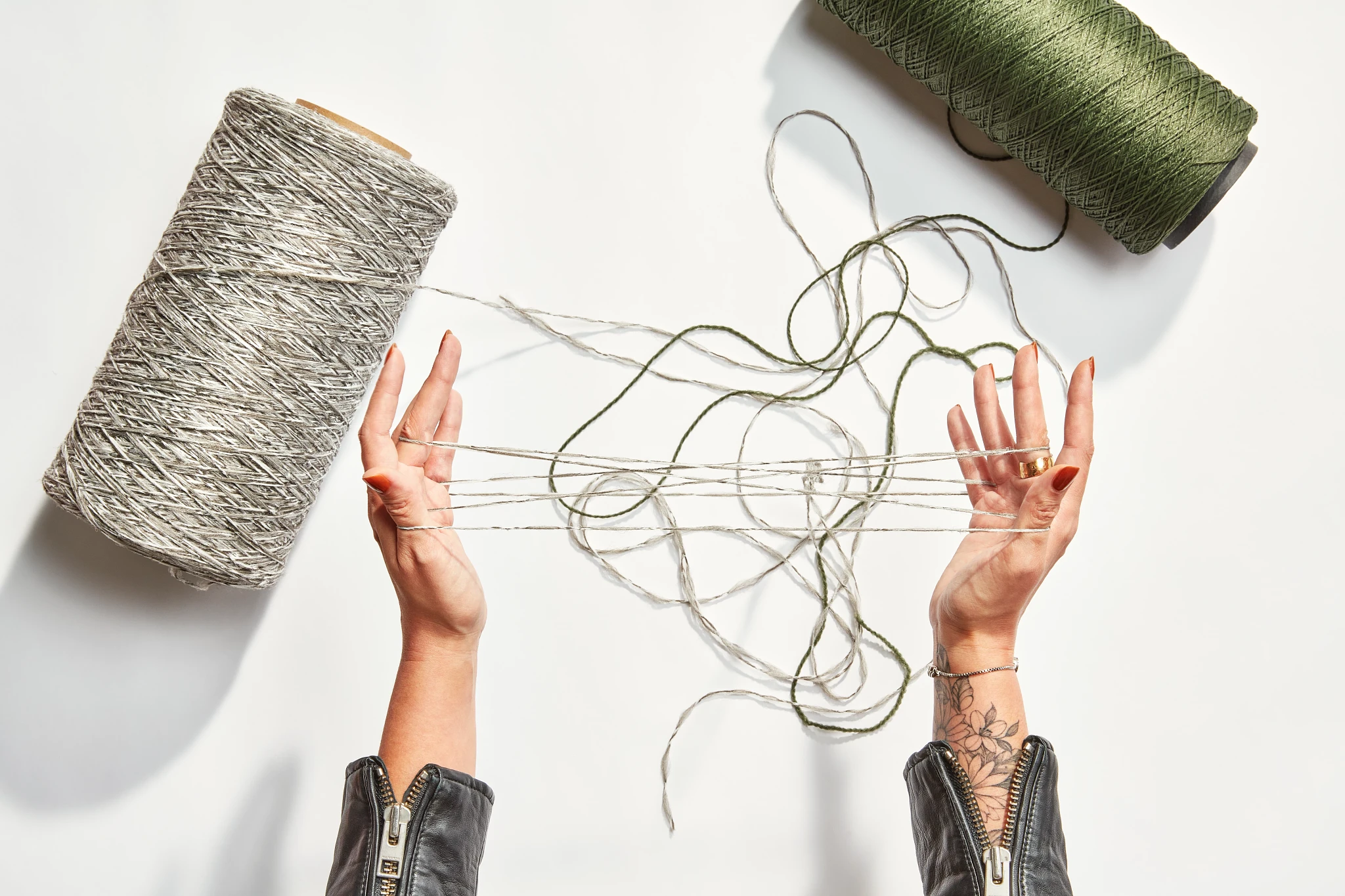 Hands holding threads with large spools of gray and green yarn on a white background