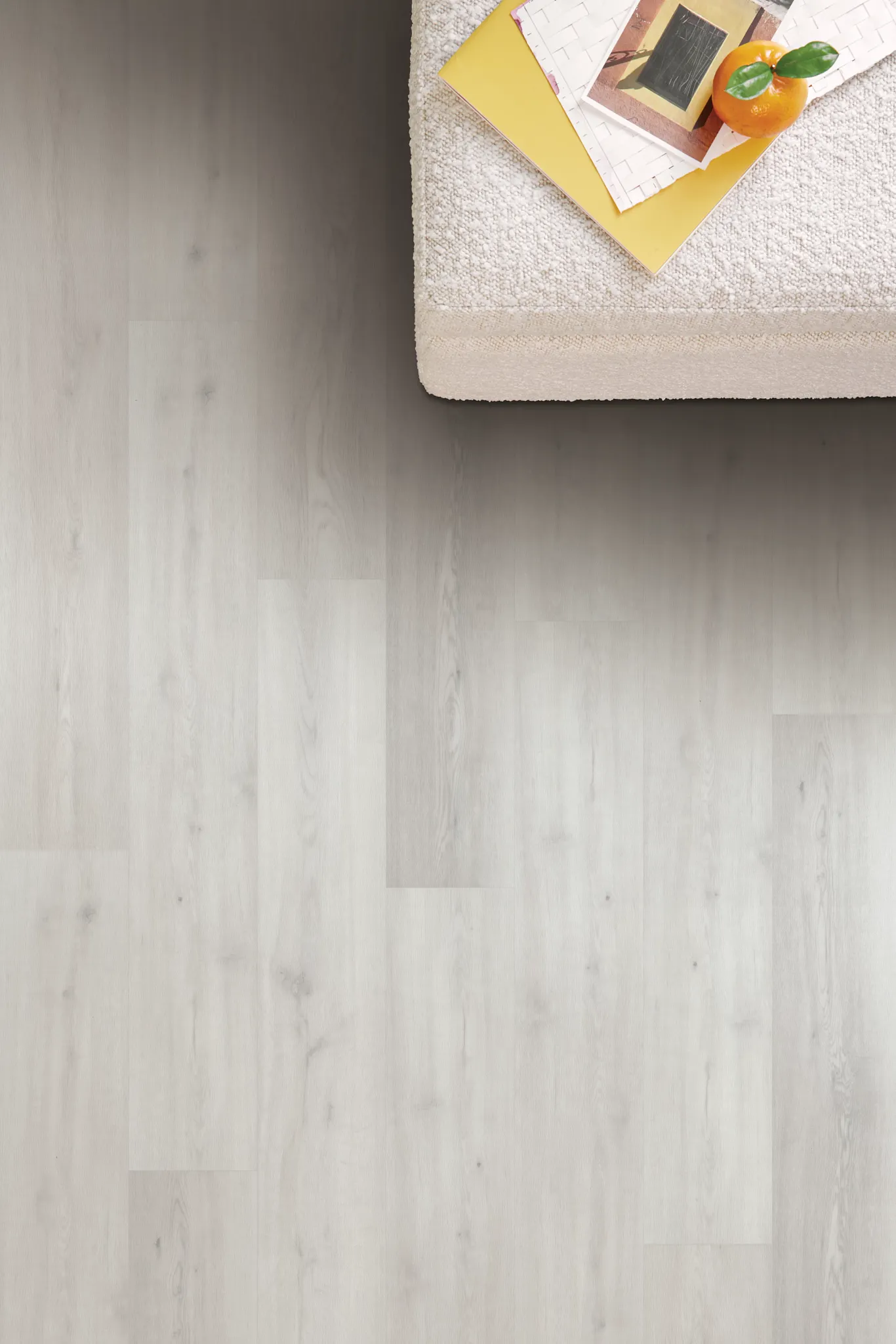 Top-down view of light wood look luxury vinyl tile flooring from the Serene collection with a textured upholstered chair and magazines in the corner.