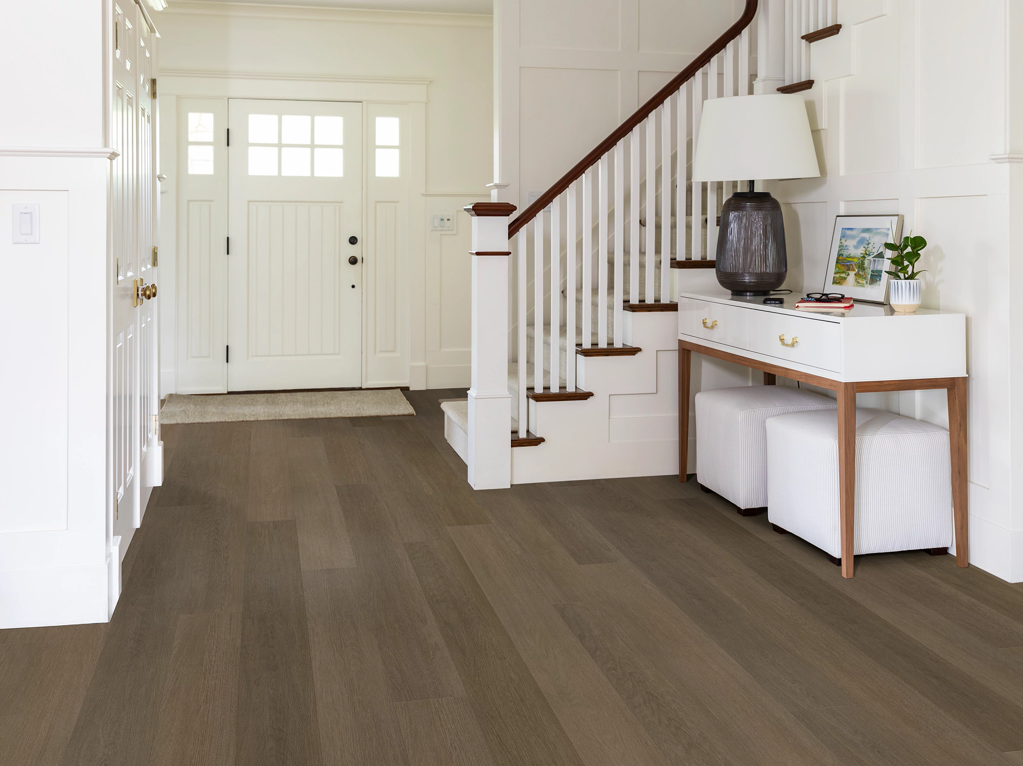 Bright entryway with white paneled walls, staircase with wooden handrail, and a console table with lamp and decor, showcasing Renegade flooring in Denali—a rich, warm brown tone with a smooth matte finish.