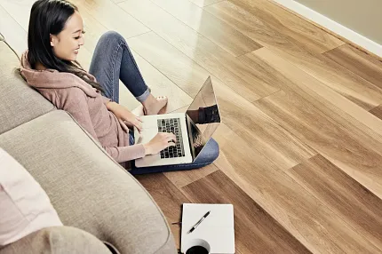SPC Flooring in Natural Wood Tones - 59.06x8.86 inch Woman working on a laptop in a cozy room with SPC flooring in natural wood tones, 59.06x8.86 inch dimensions