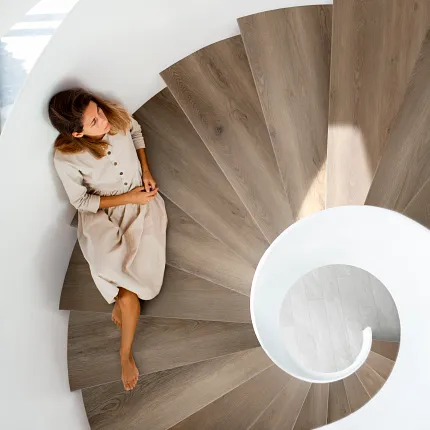 Wood-look LVP flooring on spiral staircase with modern design Spiral staircase with wood-look luxury vinyl plank flooring and a woman in a beige dress sitting on the steps