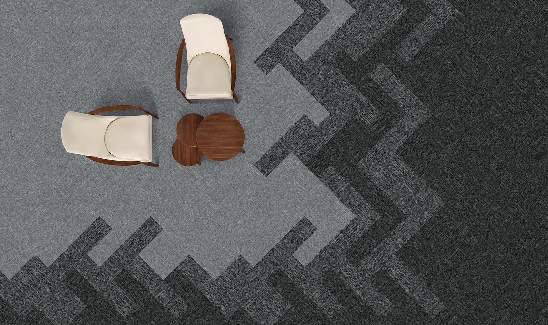 Overhead view of a modern seating area with two cream chairs and a wooden table on a patterned grey and dark grey commercial carpet tile floor
