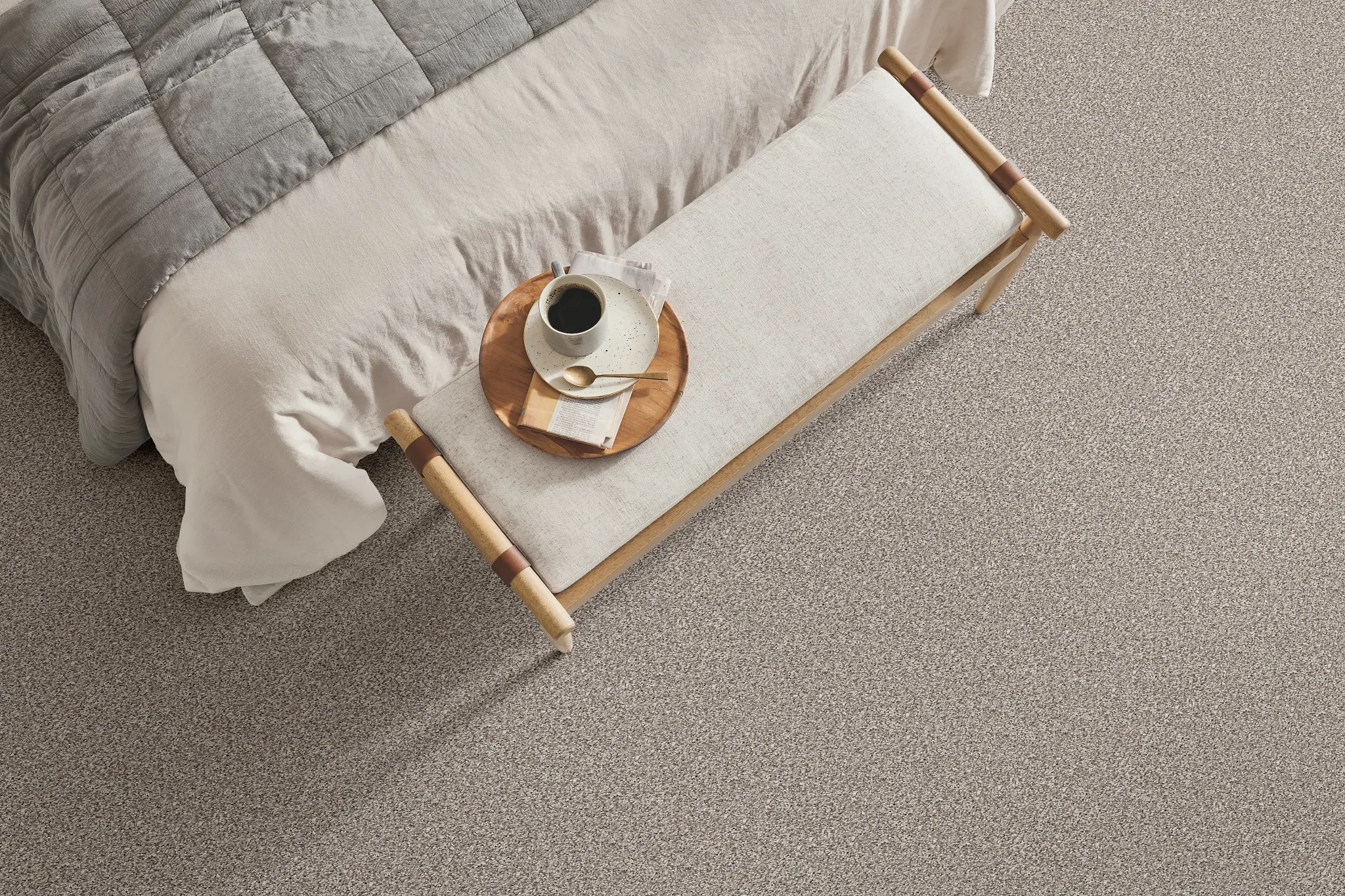 Cozy bedroom scene with a soft, textured carpet in a neutral beige tone, featuring a neatly made bed with a gray quilt and a wooden bench with a breakfast tray