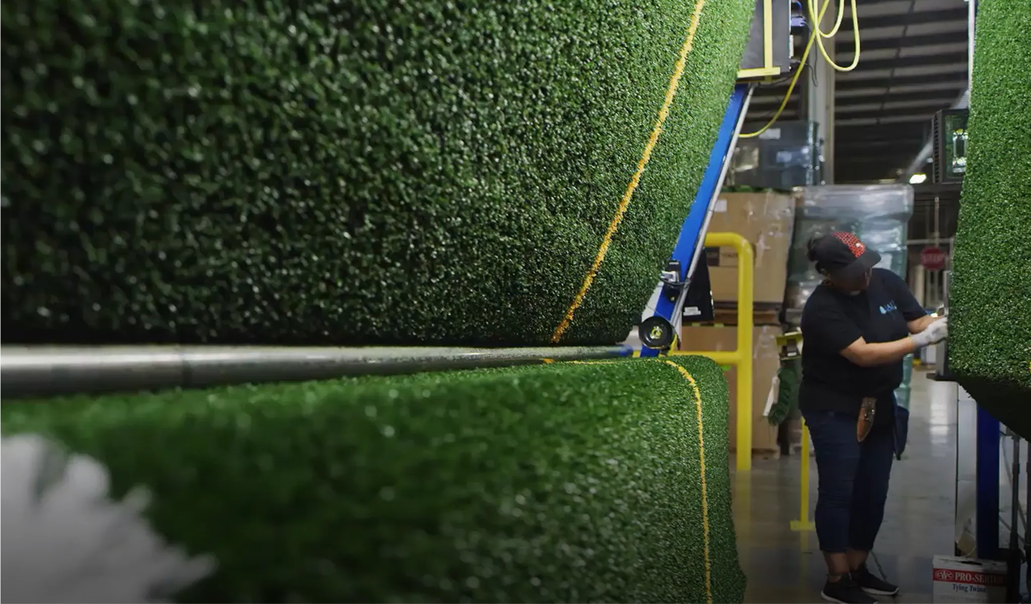 Worker inspecting large rolls of synthetic turf inside a manufacturing facility.