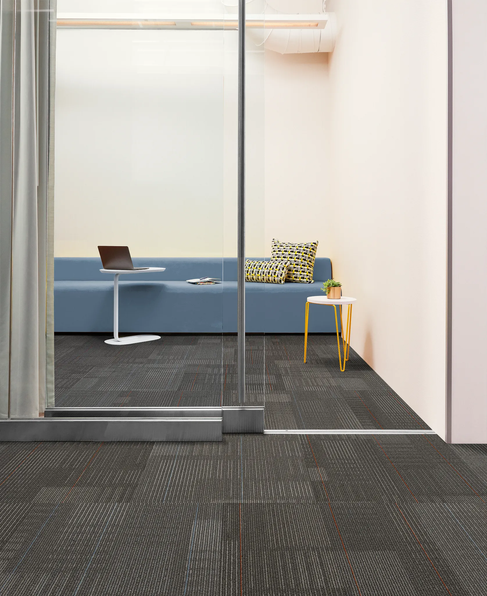 Modern office room with glass partition, featuring dark gray carpet tiles with subtle blue and red lines, blue sofa, and yellow side table