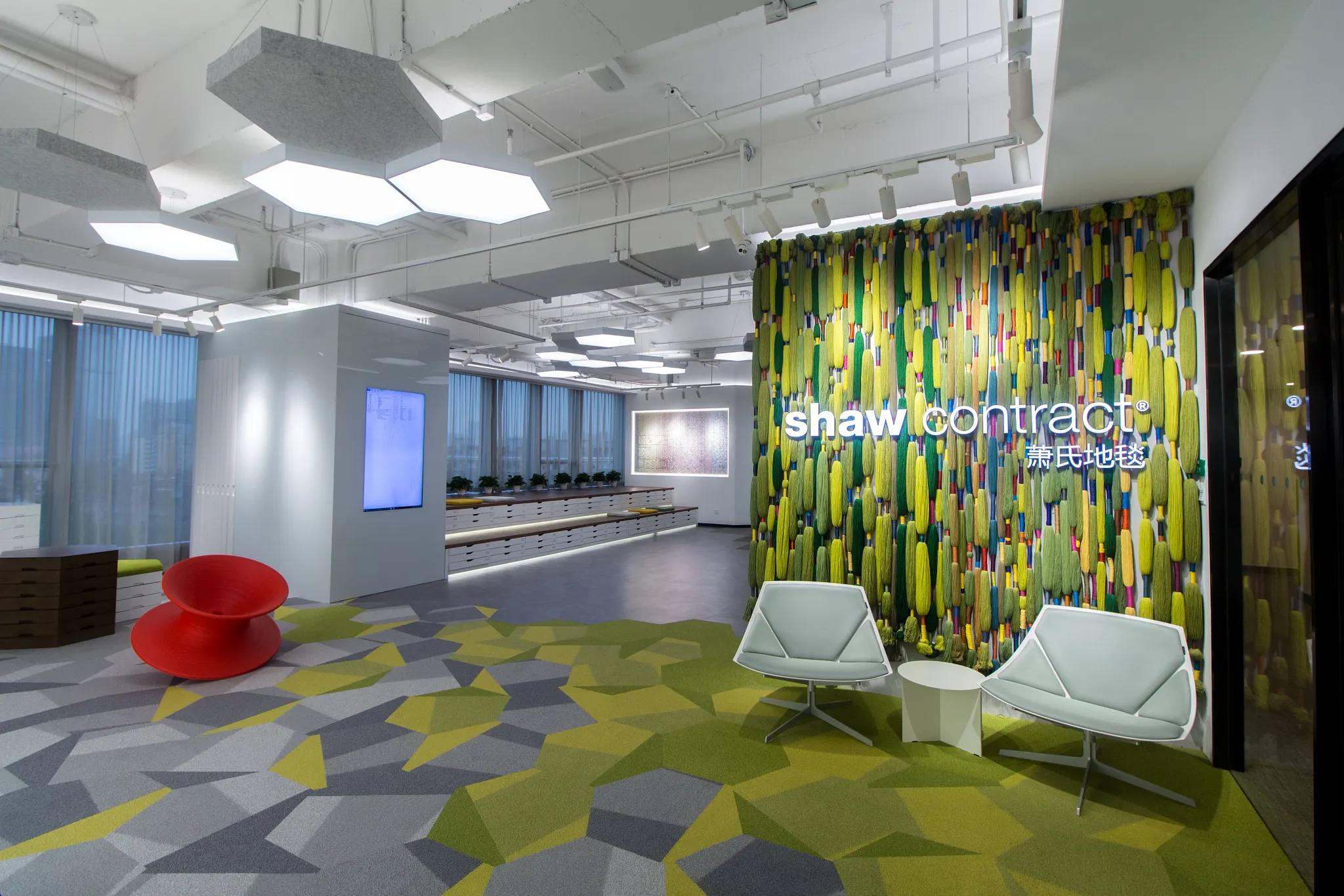 Modern office space with geometric patterned carpet in shades of green, gray, and yellow, featuring Shaw Contract branding on a vibrant wall