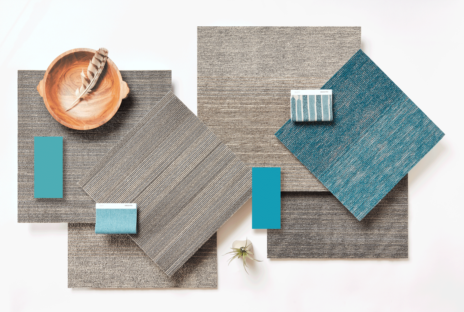 Swatch of commercial carpet tiles in shades of gray and teal with a wooden bowl and feather accent