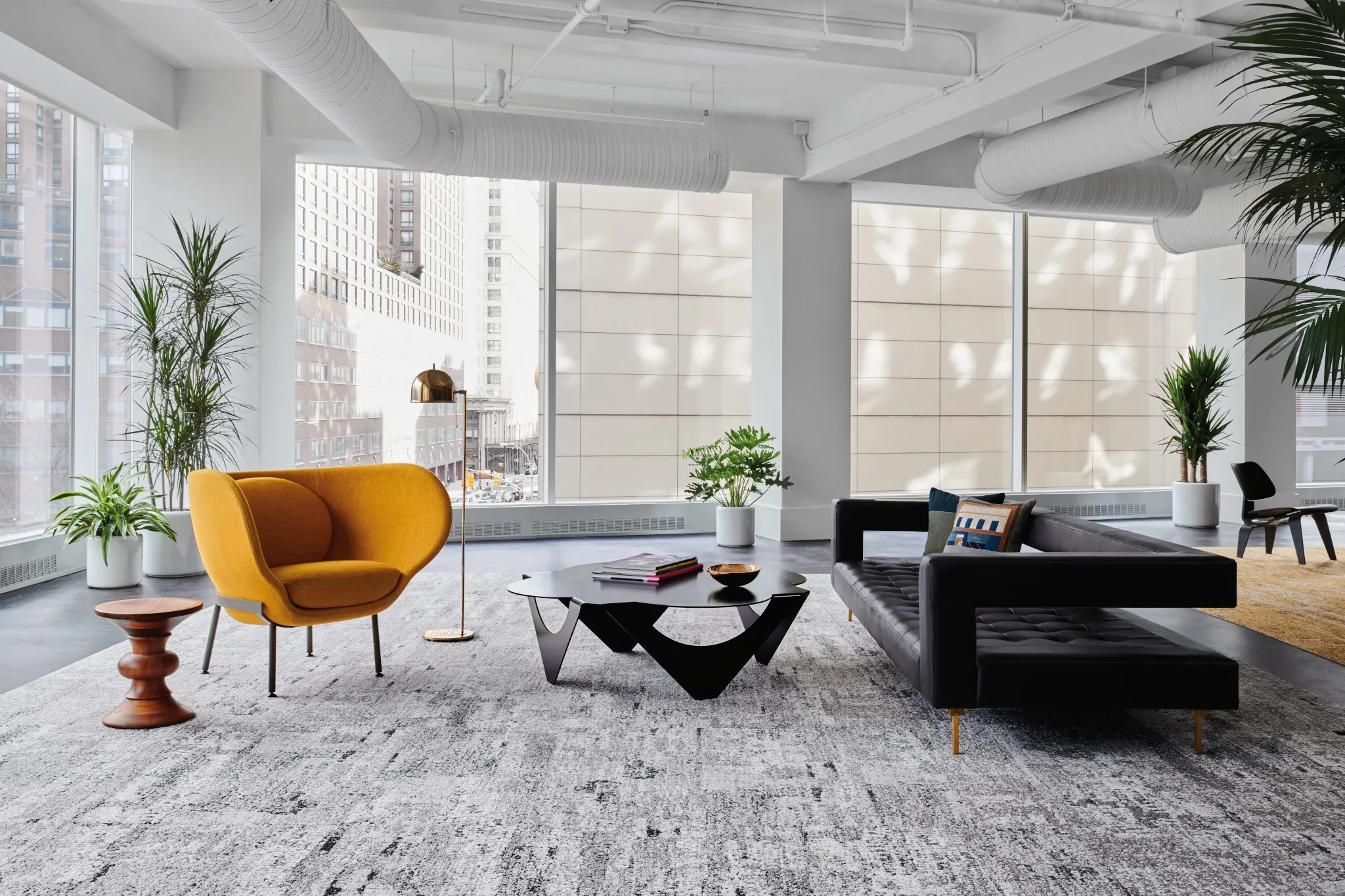 Modern office space with large windows, featuring a gray textured carpet, a yellow armchair, a black sofa, and indoor plants
