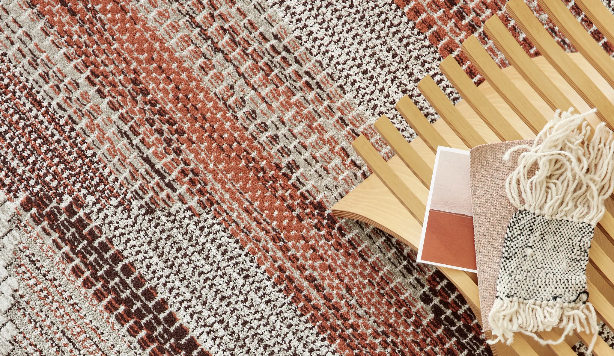 Close-up of a textured commercial carpet tile in shades of rust, brown, and cream with a wooden chair and fabric swatches