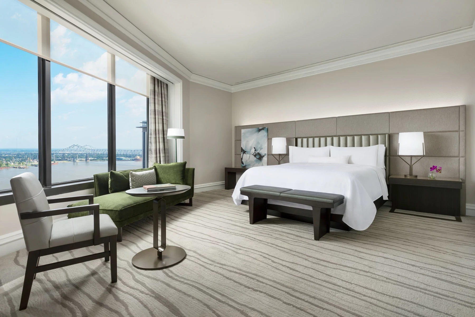 Modern hotel room with a large bed, green sofa, and a view of the river, featuring elegant striped carpet flooring, Hospitality.