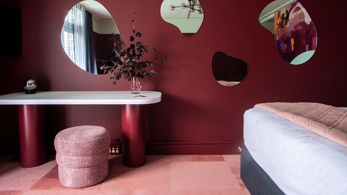 Modern hotel room with pink carpet flooring, burgundy walls, and abstract mirrors