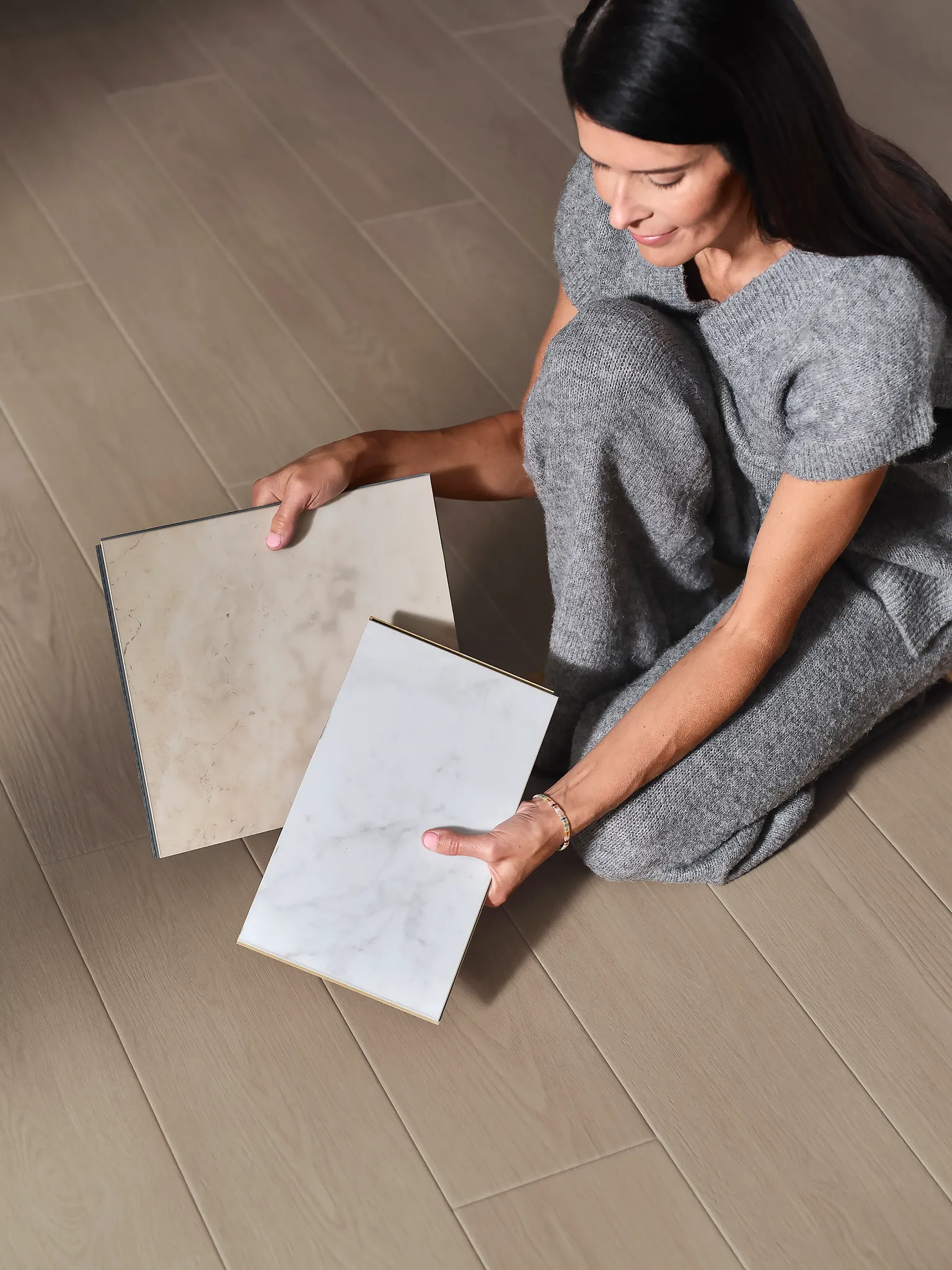 Woman in gray outfit holding two vinyl tile samples in beige and white marble patterns on wood look vinyl flooring