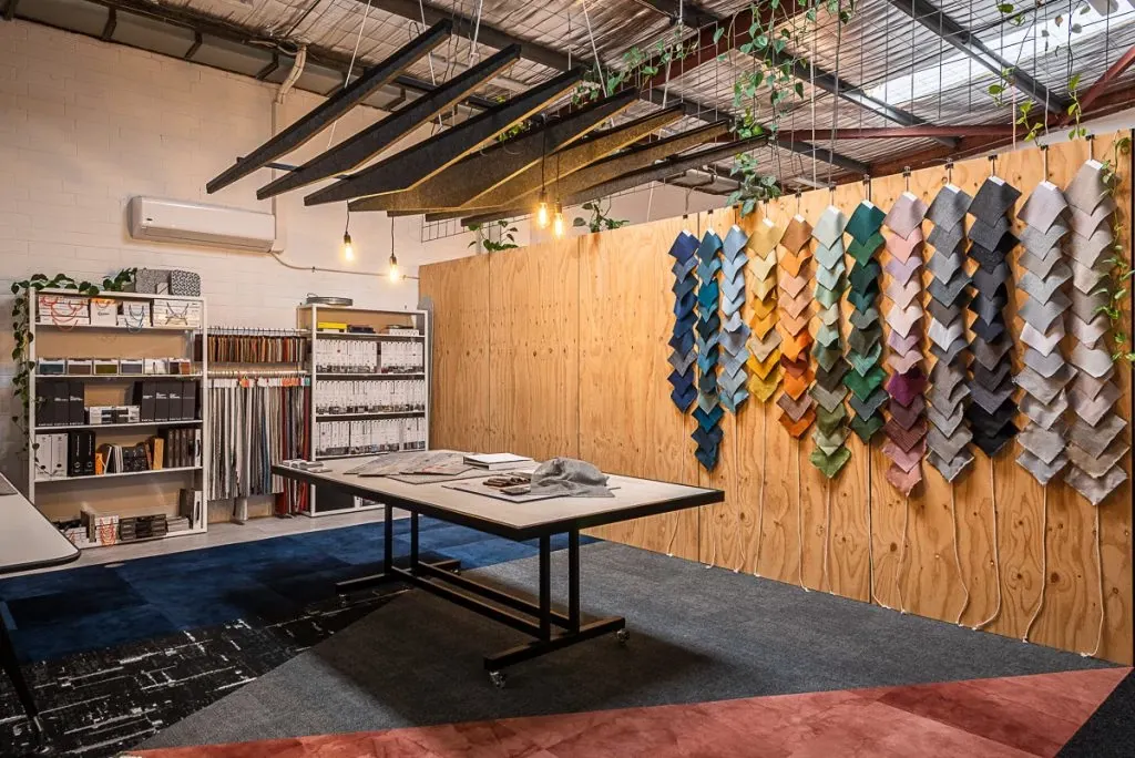 Modern showroom with a variety of colorful carpet swatches displayed on a wooden wall, featuring a mix of blue, green, orange, pink, and gray tones, with industrial lighting and shelving in the background