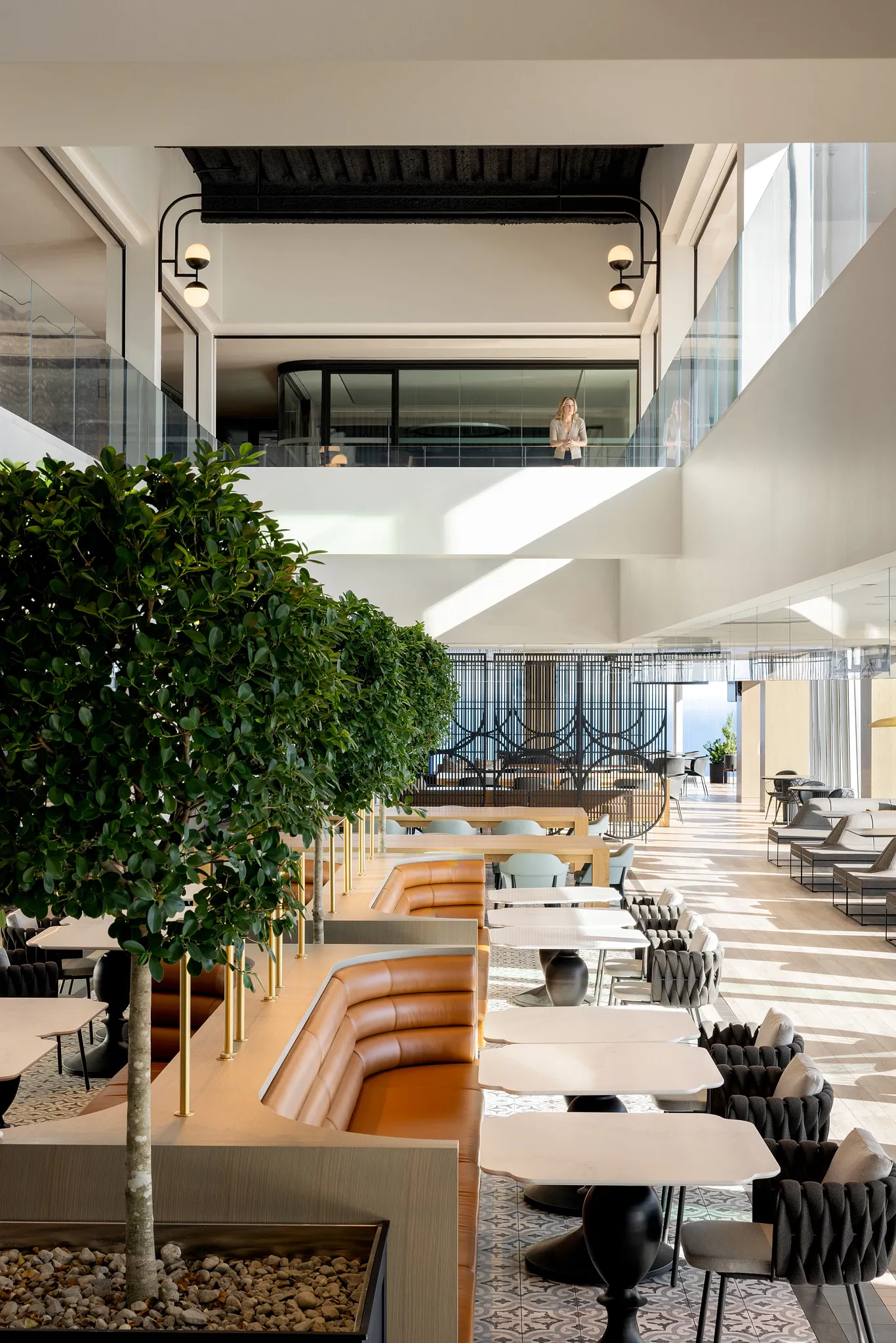 Modern workplace interior with stylish seating, large potted plants, and a woman standing on the upper level
