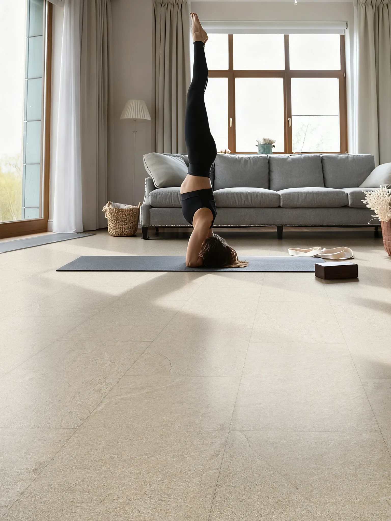 Person practicing yoga in a modern living room featuring light stone-look luxury vinyl flooring, gray sofa, large windows, and natural daylight