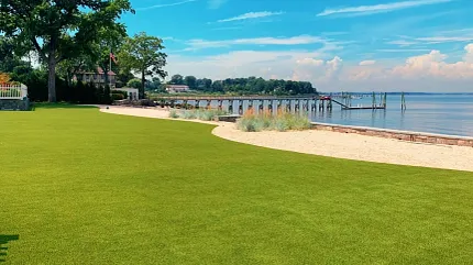 Waterfront Artificial Turf Lawn Installation | Shaw Grass Lush artificial turf lawn installed along a waterfront property with sandy landscape edging and a wooden pier in the background.
