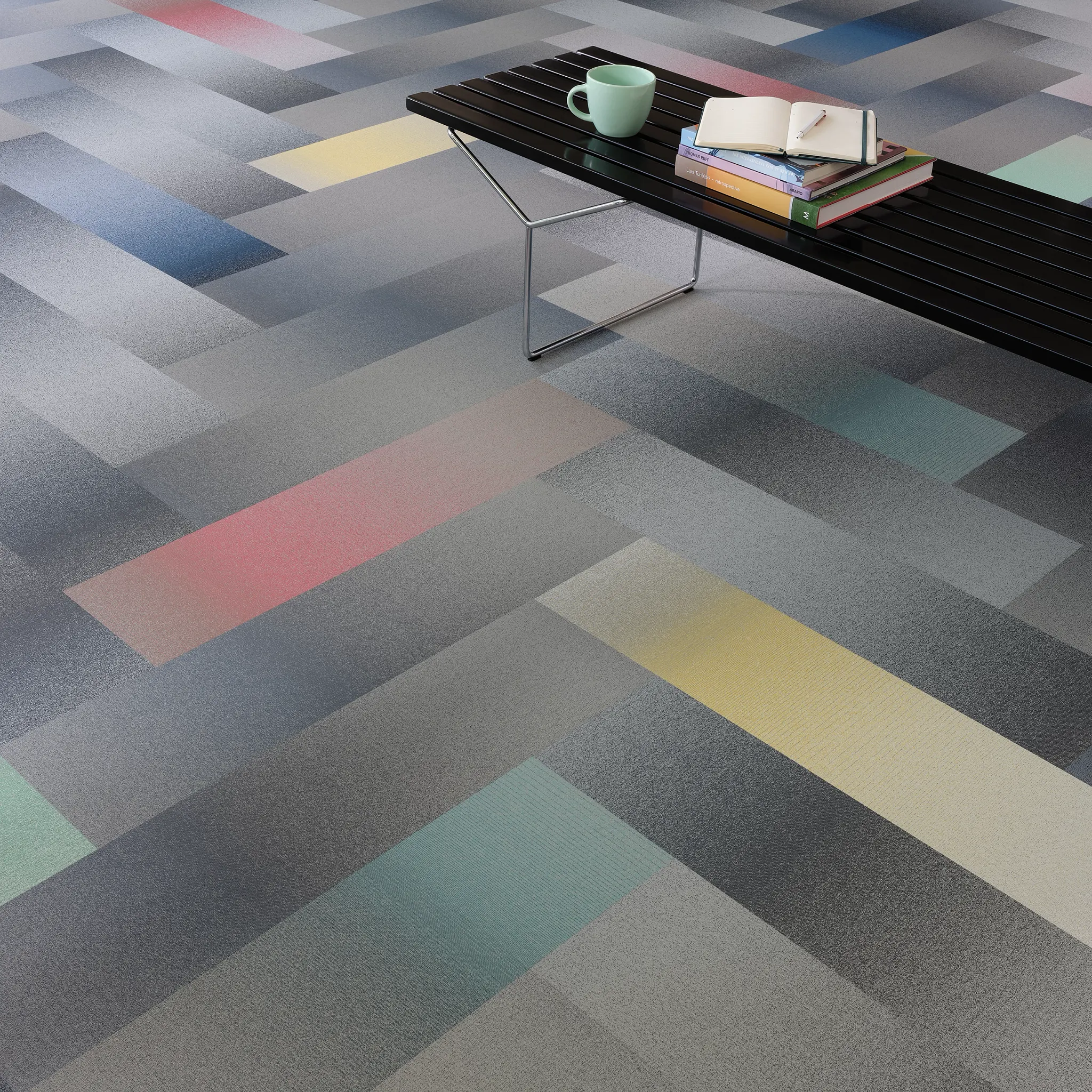 Modern commercial carpet tile in shades of gray, blue, red, yellow, and green with a black bench and books in an education facility setting