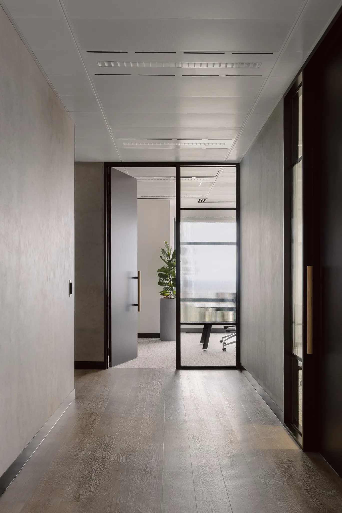 Modern office hallway with sleek hardwood flooring and glass door leading to a meeting room with a potted plant
