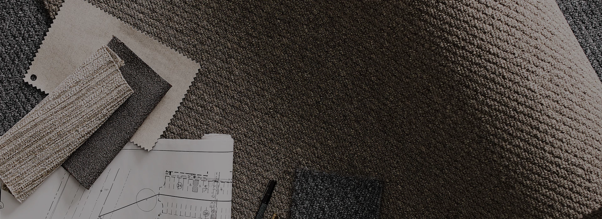 Carpet swatches in shades of beige, brown, and gray on a textured flooring surface with architectural plans and a pen