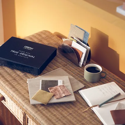 Cozy Workspace with Fabric Swatches and Coffee on Wicker Desk A cozy workspace with a wicker desk featuring a black box labeled "From One True Original to Another," fabric swatches in various colors, a cup of coffee, and a notebook with a pen