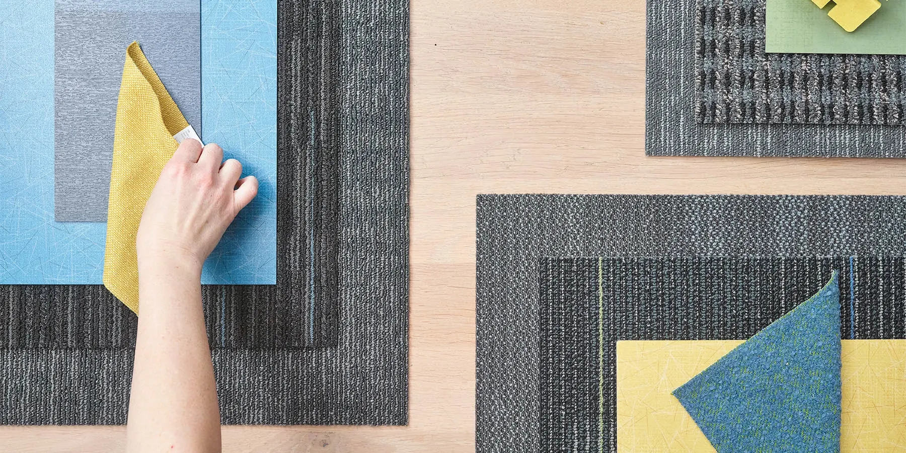 Hand arranging commercial carpet tile samples in blue, yellow, and gray on a wooden surface