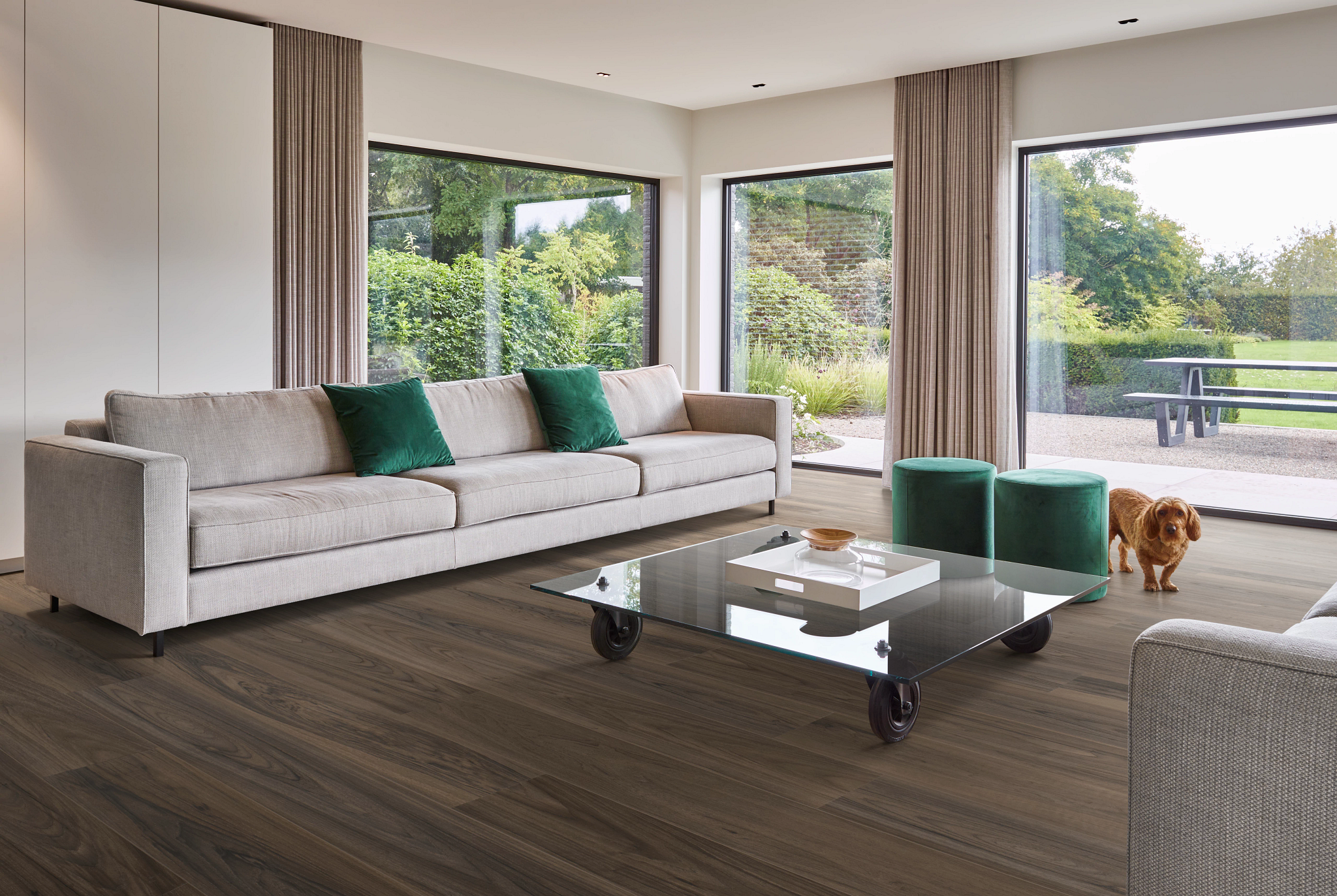 Modern living room with large windows, beige sofa, green cushions, and a dog on dark wood look luxur…