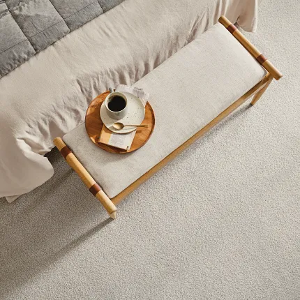 Beige Carpet Flooring in Cozy Bedroom Scene with Gray Quilt and Wooden Bench Cozy bedroom scene with soft beige carpet flooring, featuring a neatly made bed with a gray quilt and a wooden bench with a coffee cup and saucer.