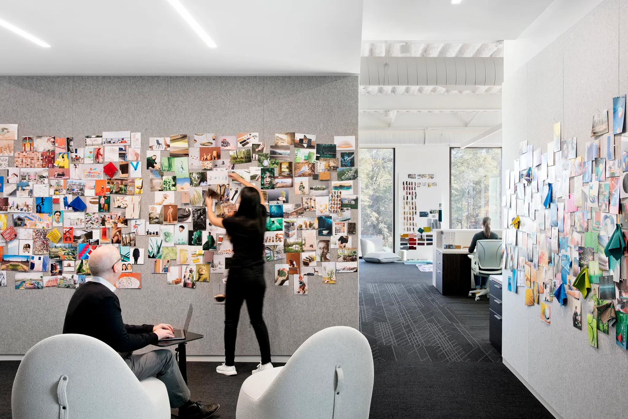 Modern office space with two people collaborating, featuring dark carpet flooring and creative wall displays