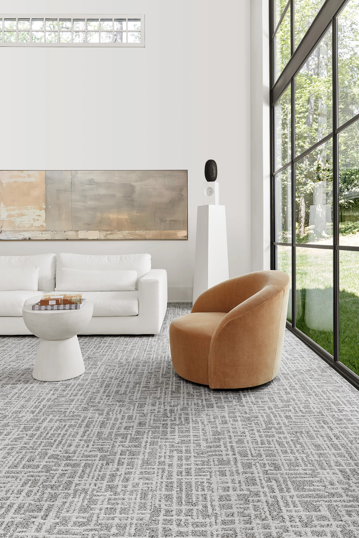 Modern living room with large windows, featuring a white sofa, beige armchair, and abstract art on the wall, with a patterned gray carpet flooring