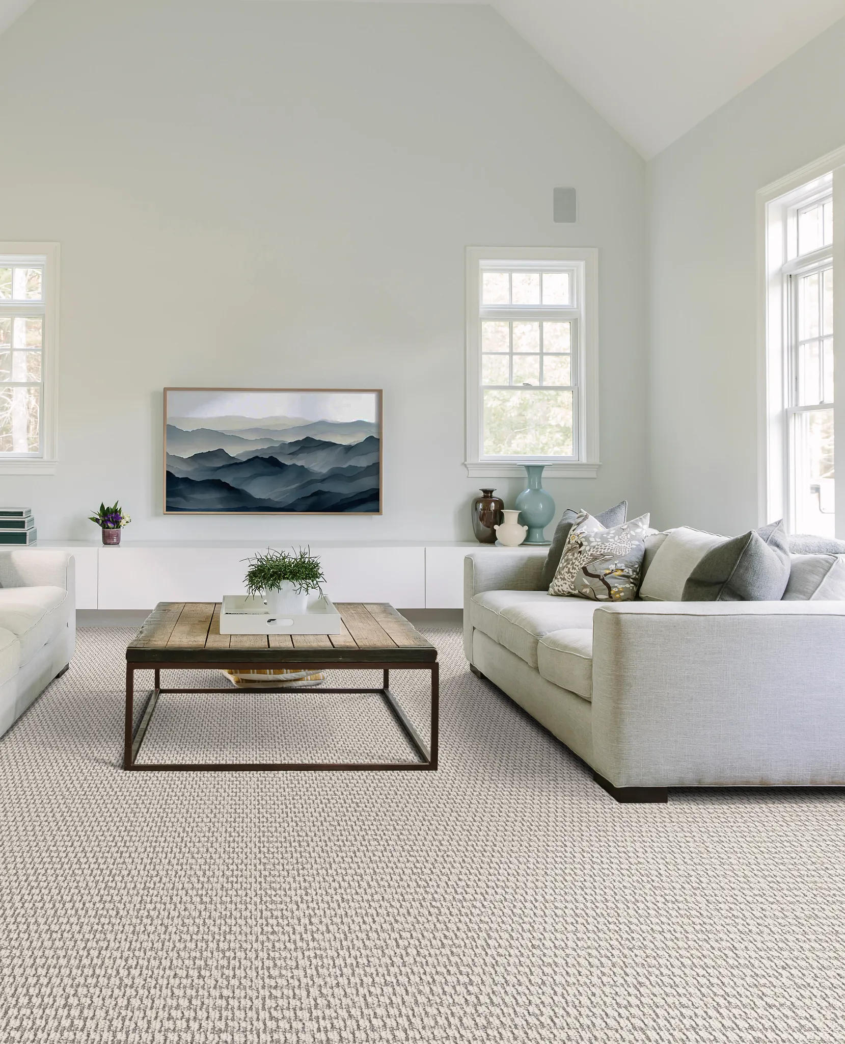 Modern living room with soft surface flooring, featuring a beige and gray patterned carpet, two light gray sofas, a wooden coffee table, and a landscape painting on the wall