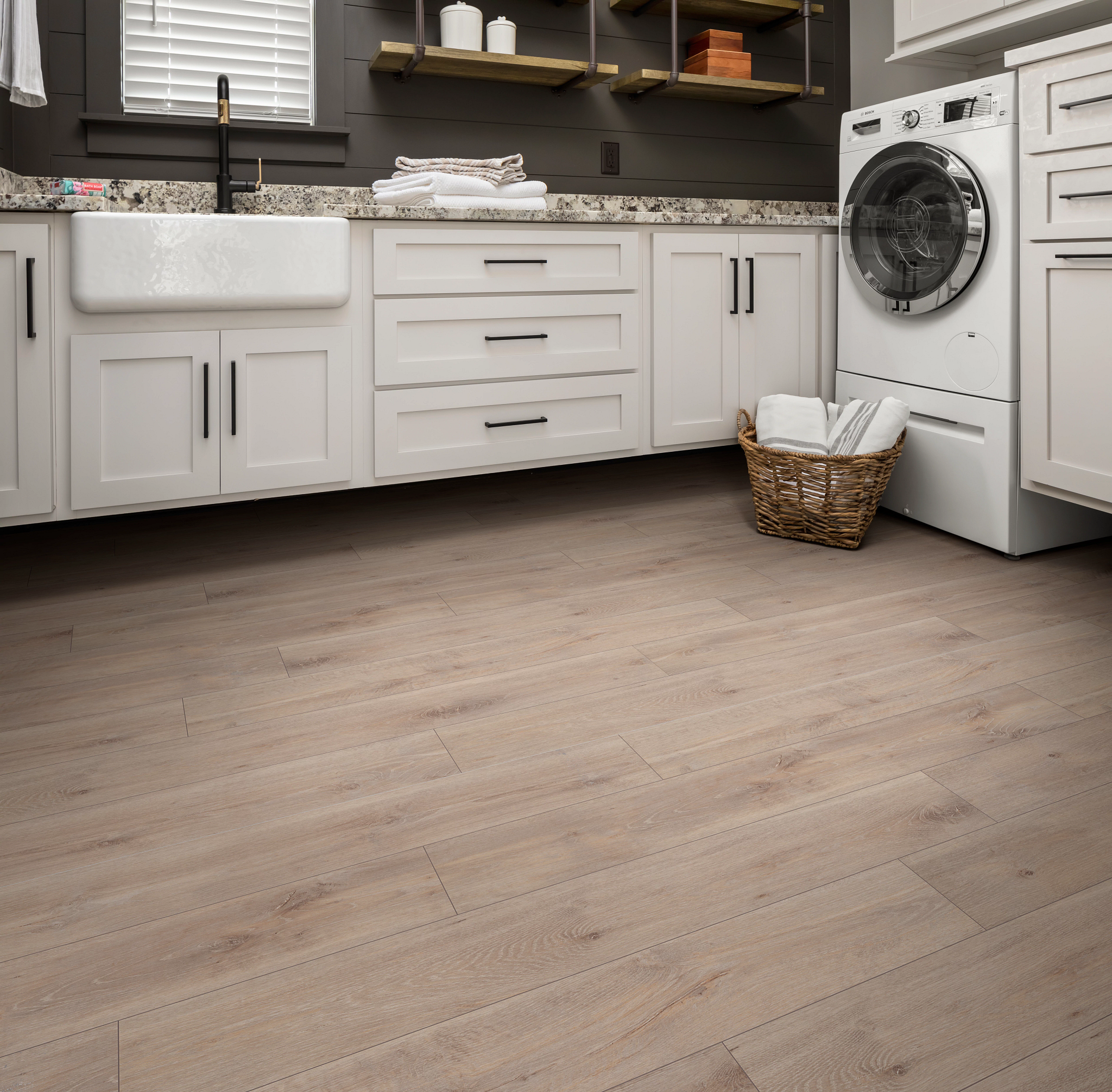 Laundry room with white shaker cabinets, farmhouse sink, granite countertops, and front-load washer, featuring Pride Plus Accent SPC flooring in Ryman with soft beige wood tones and a matte finish.